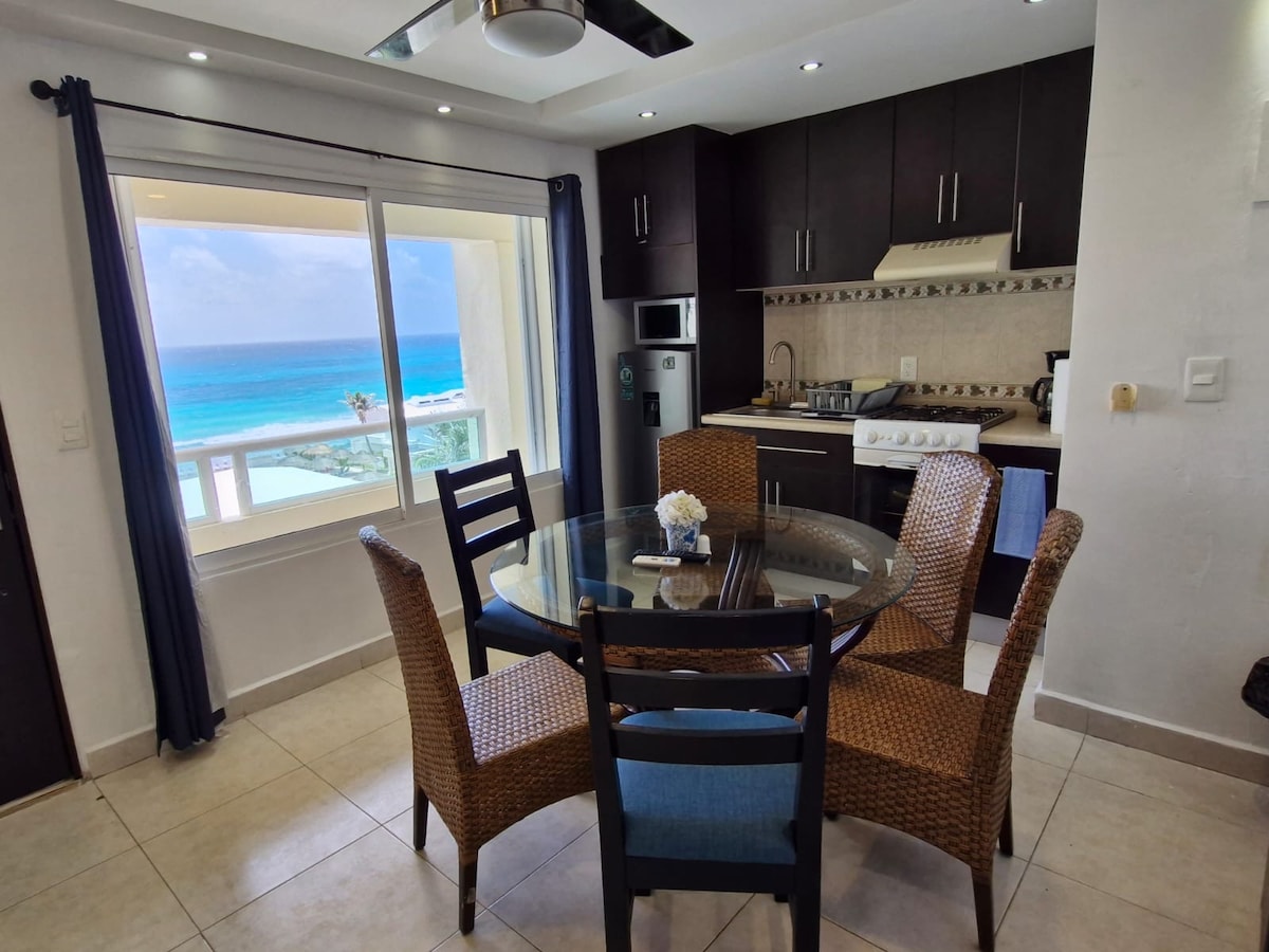 A dining area is set with a glass table surrounded by four woven chairs, placed near a window that offers a clear view of the ocean. A kitchenette is visible in the background, equipped with essential appliances and dark cabinetry.