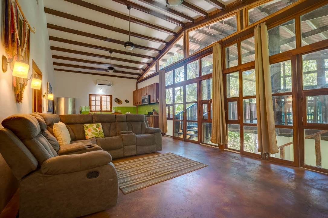 A spacious living area features an inviting sectional sofa positioned along a textured floor. Tall windows with wooden frames allow natural light to fill the space, highlighting the open ceilings with beams. Curtains frame the windows, complementing the relaxed atmosphere of the room.