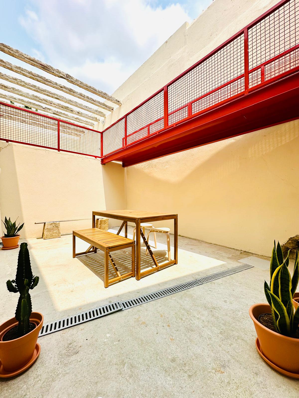 An open courtyard features a wooden dining table and chairs under a retractable roof. Green potted plants are positioned in each corner, adding a touch of nature. Natural light illuminates the space, highlighting the simplicity and functionality of the design.