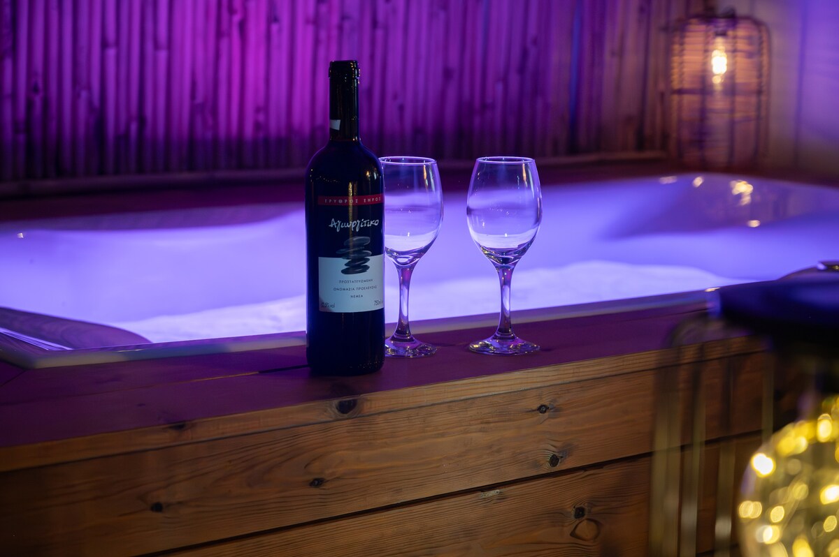 A wooden table is set beside a jacuzzi, featuring a bottle of red wine and two empty wine glasses. Soft purple lighting enhances the ambiance, with bamboo accents visible in the background, creating a relaxing environment.