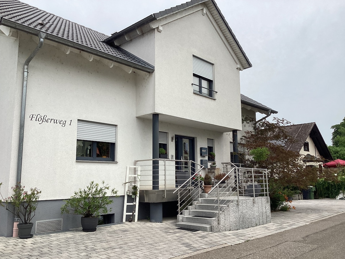 A two-story building is viewed from the front, featuring a light-colored exterior and a covered entrance with steps. The walkway is lined with decorative plants, and a railing enhances accessibility. Windows allow natural light, framing a simple and functional design.