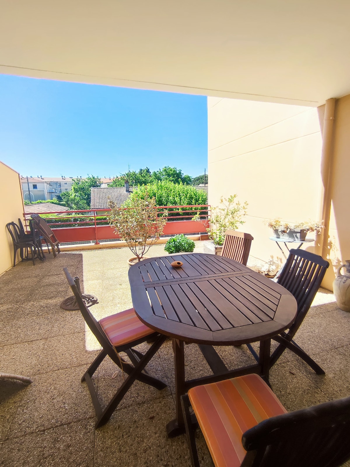 A sunlit terrace features a wooden dining table surrounded by four chairs. Potted plants add natural elements to the space, which opens to views of the greenery and buildings beyond. The walls provide a sense of privacy, enhancing the outdoor dining experience.
