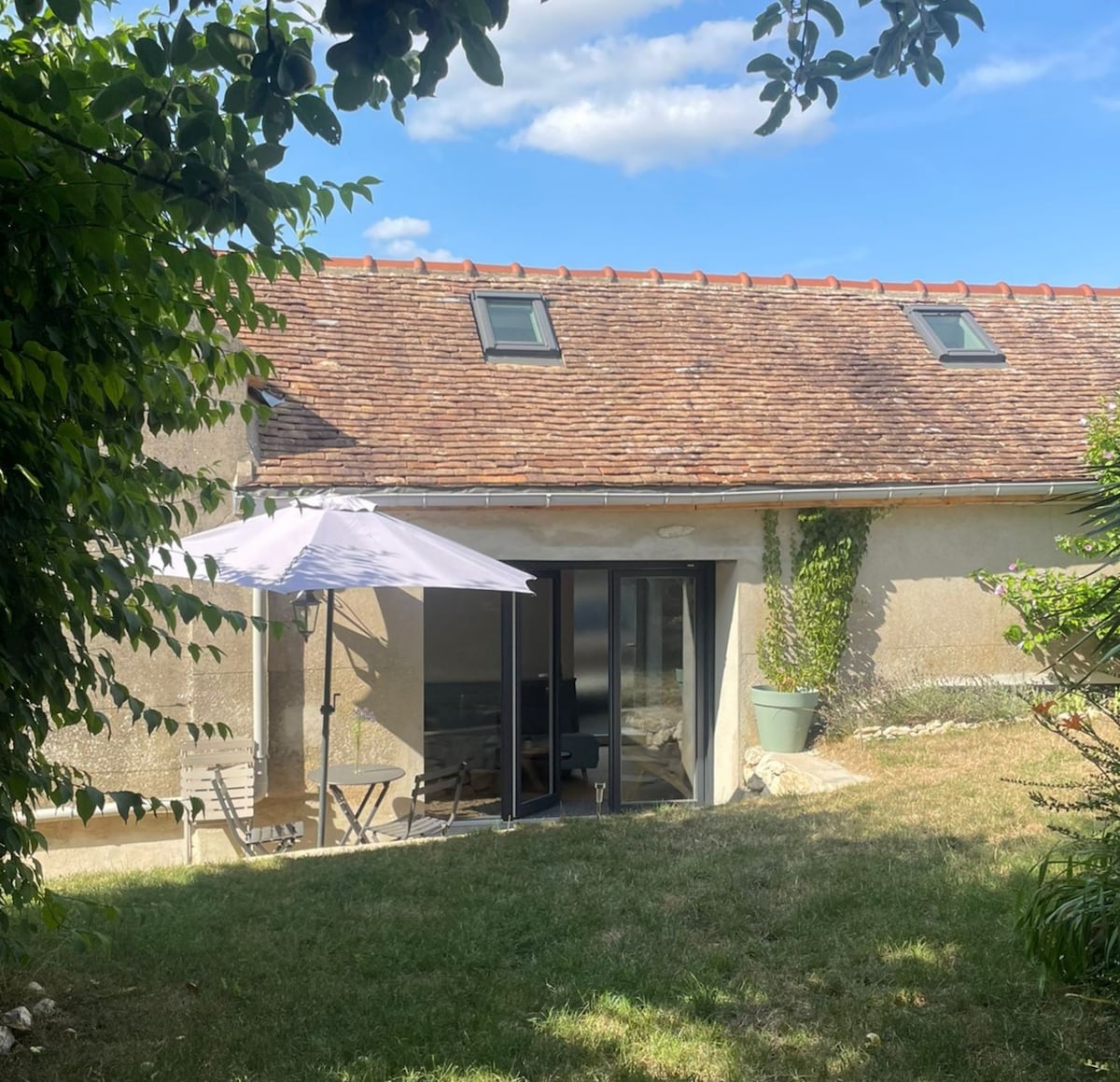 The exterior of the studio is revealed, featuring a sloping roof and large glass doors that open to a private garden space. A patio umbrella shades a small table and chairs, surrounded by greenery and a well-maintained lawn, under a clear blue sky.