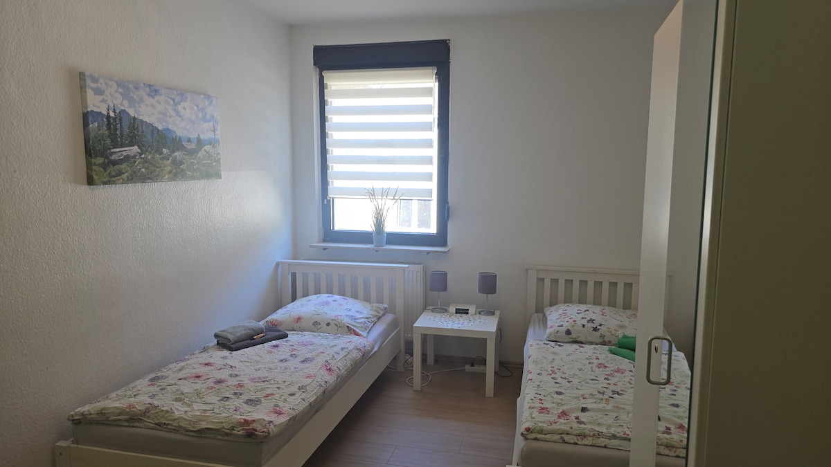 acogedor dormitorio con 2 camas individuales, un armario con cerradura y una zona de estar con 2 sillas y un televisor inteligente