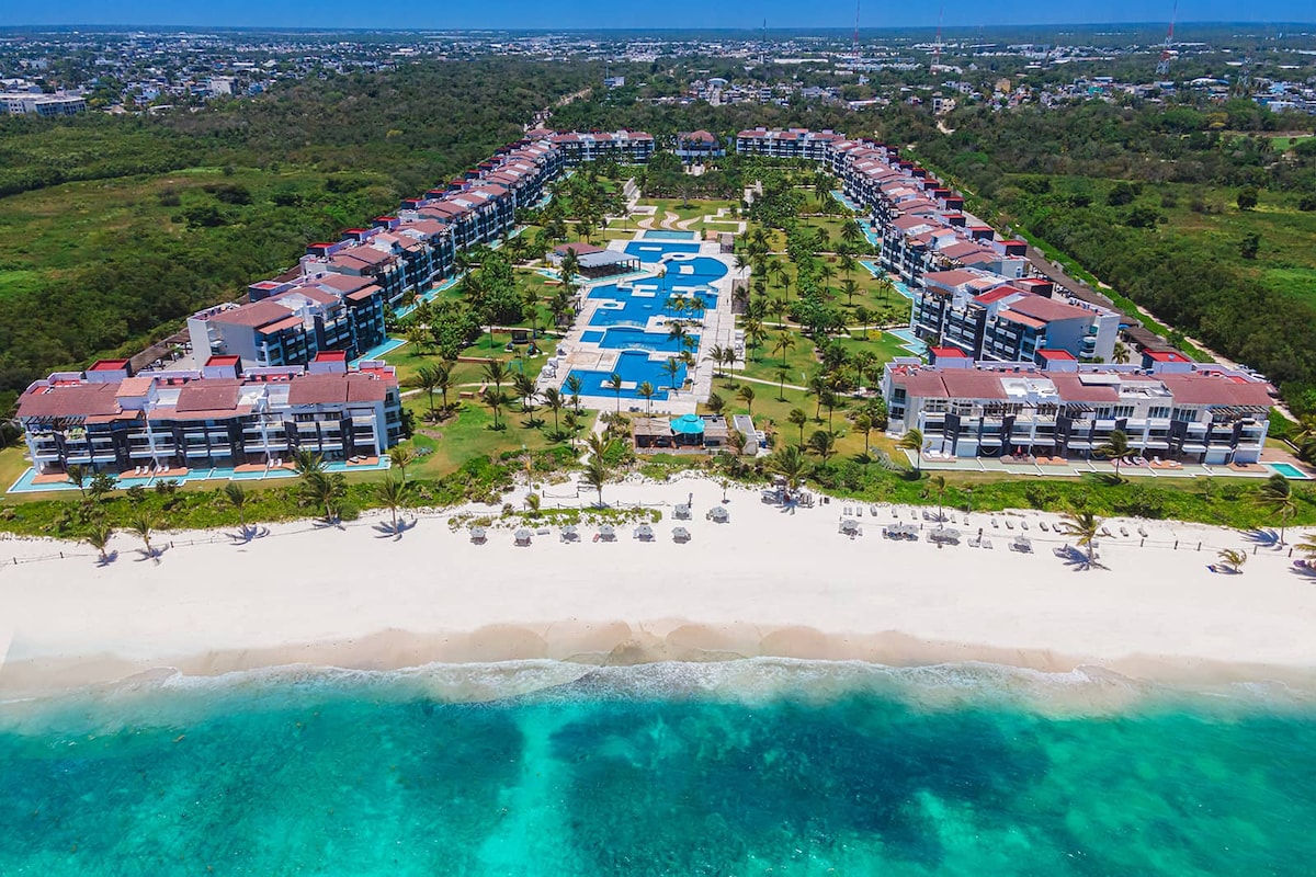 Aerial view captures a sprawling resort complex set along a sandy beach. Multiple buildings with red roofs are surrounded by lush greenery and a large swimming pool, with lounge chairs neatly arranged by the water’s edge and an inviting shoreline stretching below.