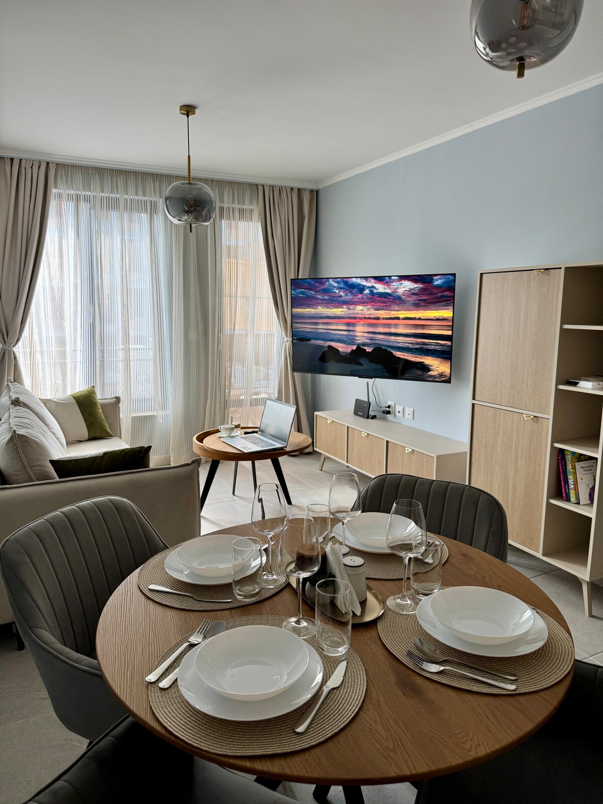 A dining area features a round wooden table set for four with dishware and glassware. A comfortable seating area includes a soft sofa positioned next to a large window, allowing natural light to fill the space. A wall-mounted TV displays a colorful ocean scene.