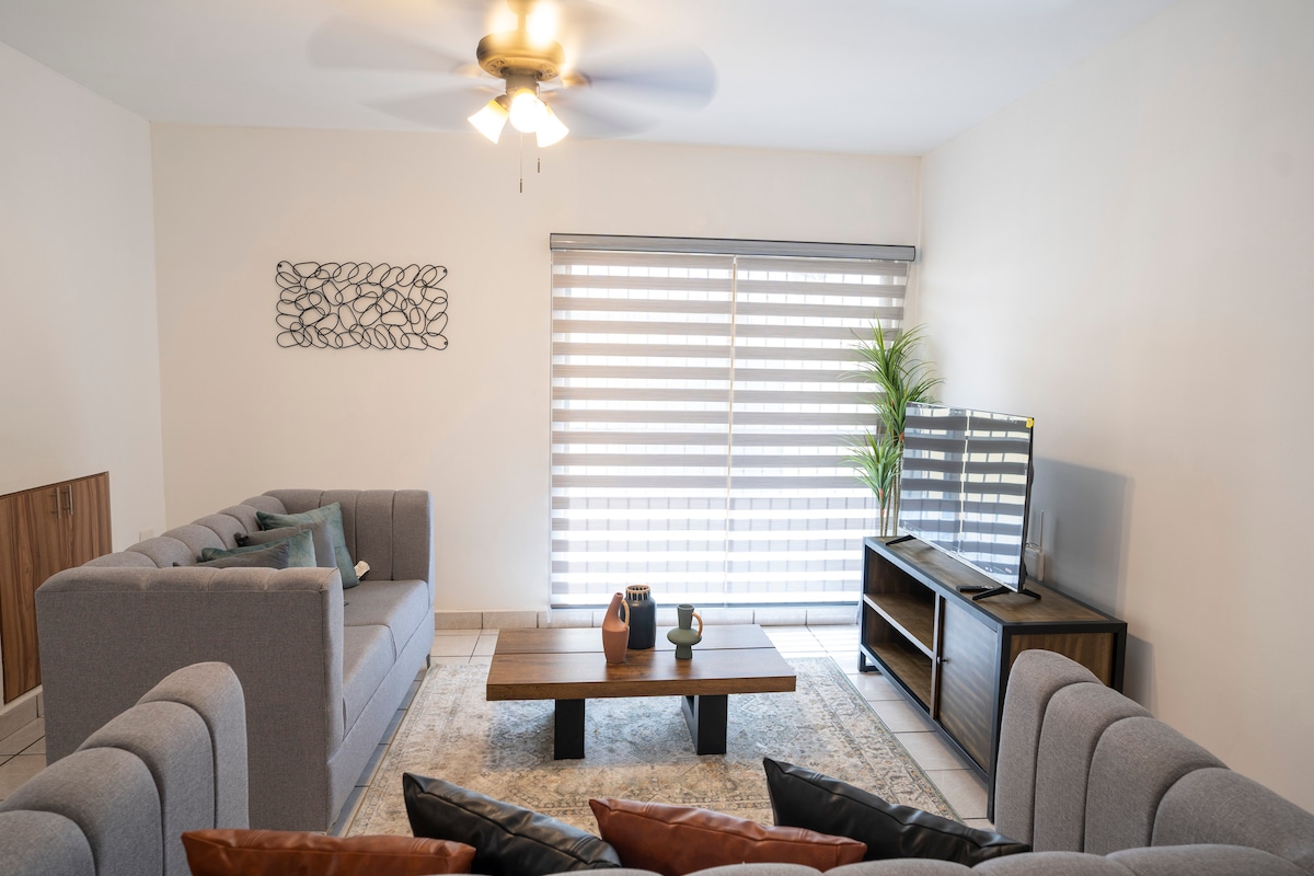 A spacious living area features two comfortable sofas and a central coffee table adorned with decorative items. Large windows with horizontal blinds provide natural light, and a television is positioned on a stand against the wall. A decorative wall piece enhances the modern aesthetic.