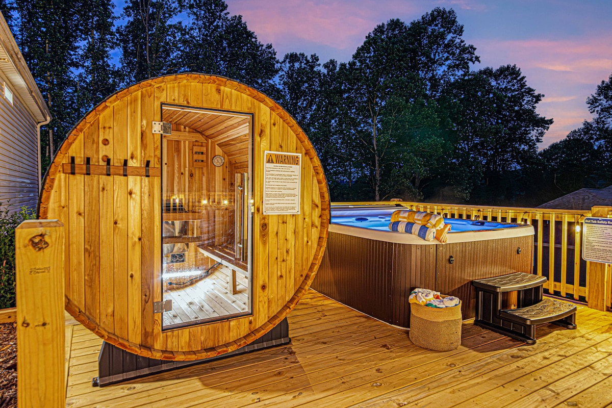 A barrel sauna made of natural wood offers a unique, traditional design, inviting relaxation with soft ambient lighting inside. Nearby, a hot tub is filled with water and features comfortable seating, set against a backdrop of lush trees and a colorful sunset sky.