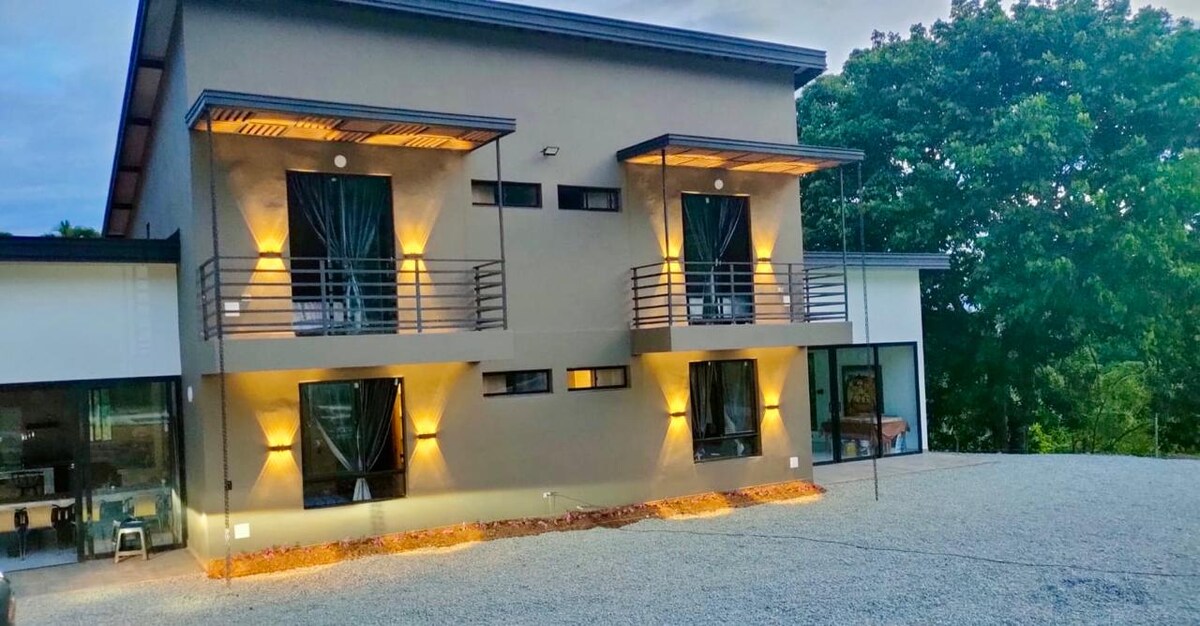 The exterior of the property features a modern two-story structure with warm outdoor lighting. Two balconies are visible on the upper level, each adorned with dark drapes, offering views of the surrounding greenery. A gravel pathway leads to the entrance, enhancing the tranquil atmosphere.