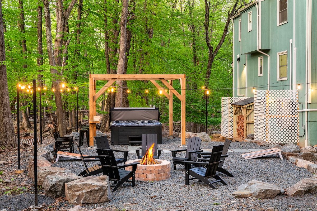 An outdoor area is highlighted by a fire pit surrounded by black Adirondack chairs. A hot tub is positioned under a wooden gazebo, with string lights draping overhead. The setting is bordered by trees, creating a peaceful retreat in a gravel landscape.