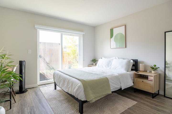 Svetla spalnica z zakonsko posteljo (Queen), sodobnim črnim okvirjem in nočnimi omaricami iz ratana z vrati za polnjenje USB. Urejena zelena dekoracija, delovna miza in drsna vrata do zasebnega zunanjega zelenega območja. Popoln oddih z naravno svetlobo!