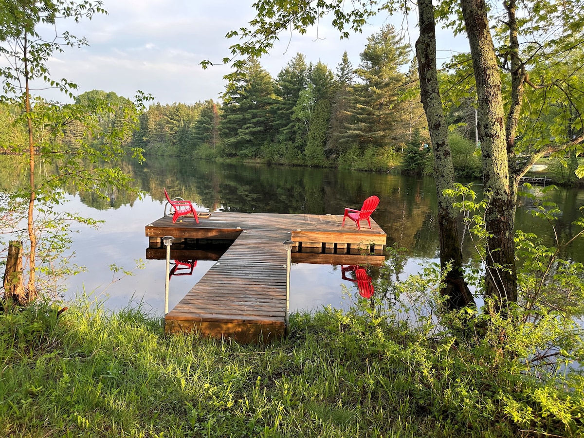 A wooden floating dock extends over calm waters, surrounded by lush greenery. Two red Muskoka chairs are positioned at the edge of the dock, inviting relaxation with serene river views and reflections from the trees along the water's edge.