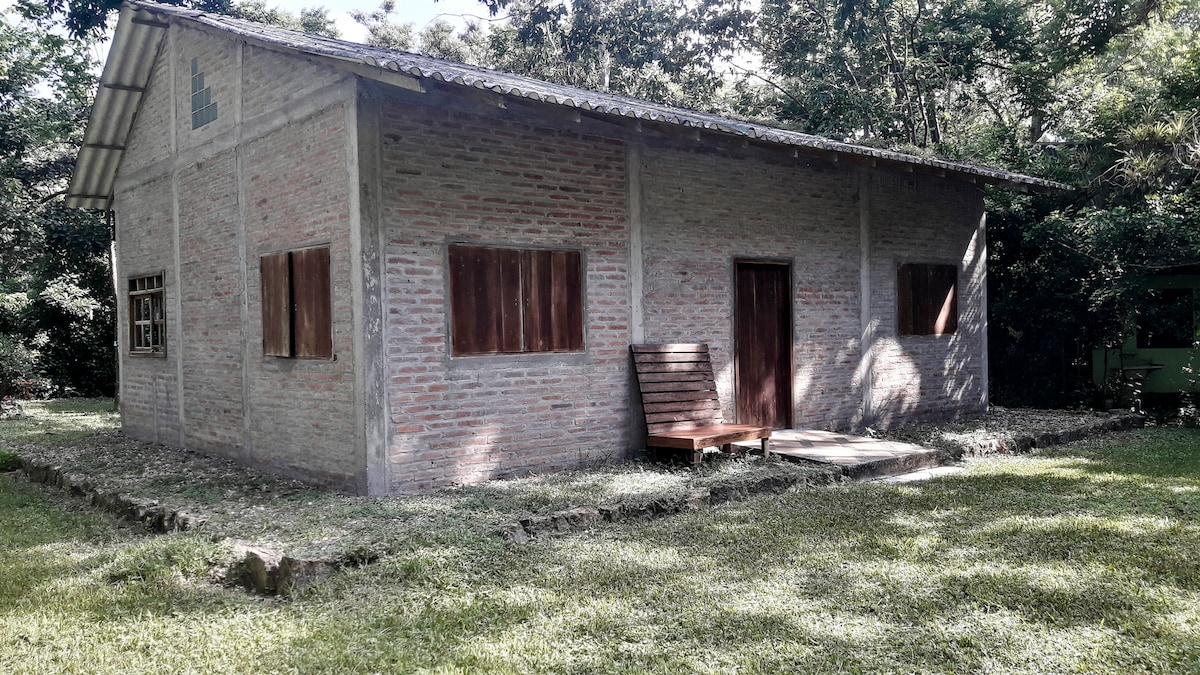 A rustic house made of brick and wood is surrounded by a lush green garden. Natural light illuminates the exterior, highlighting the wooden door and windows. A wooden bench is placed on the porch, offering a quiet spot to enjoy the outdoor space.