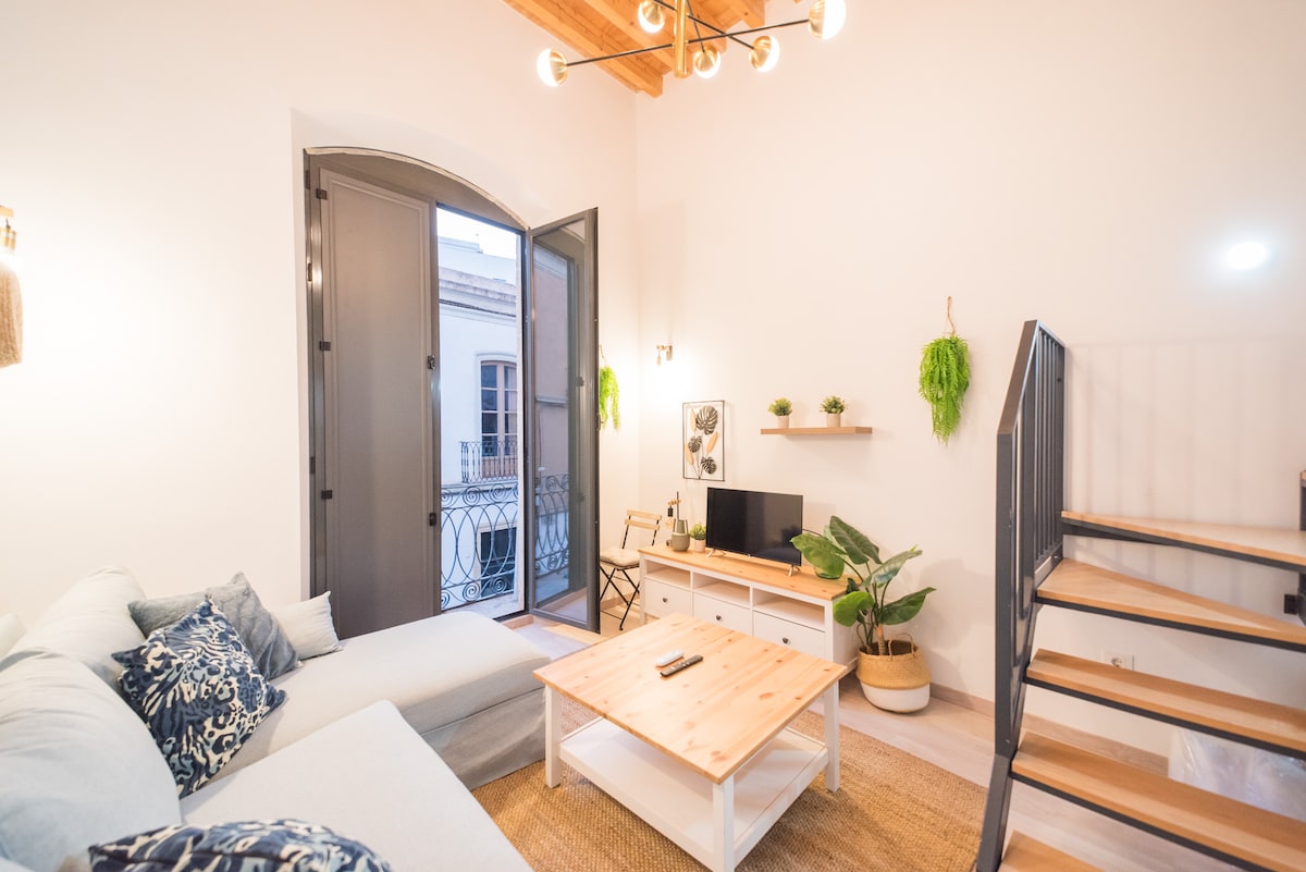 A bright living area features a light-colored sectional sofa adorned with decorative pillows. A wooden coffee table is centered on a natural fiber rug. Large, double doors open to a small balcony, while a staircase leads to the upper level. Green plants and a television enhance the space.