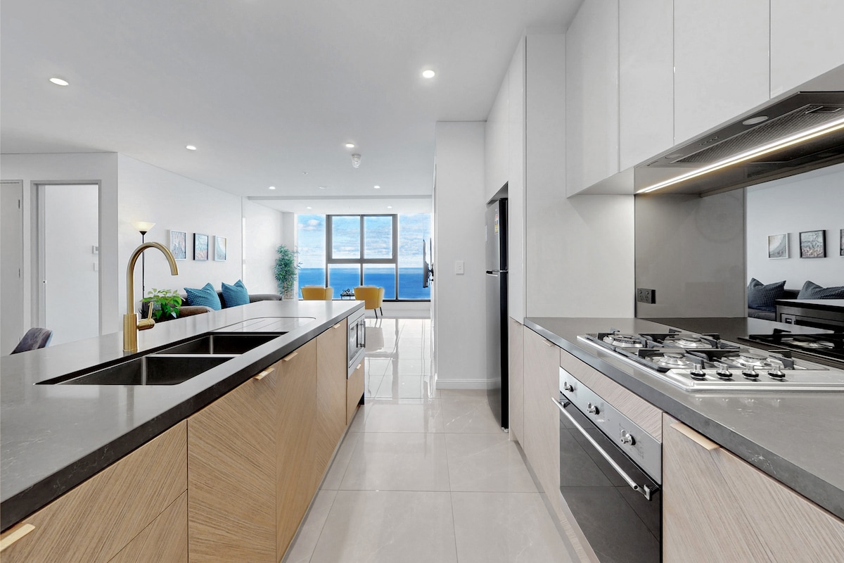 A spacious kitchen features modern cabinetry with a blend of wood and sleek finishes. A large island with a dual sink offers ample preparation space. Natural light floods the area, illuminating the seamless transition to the adjacent living space with ocean views.