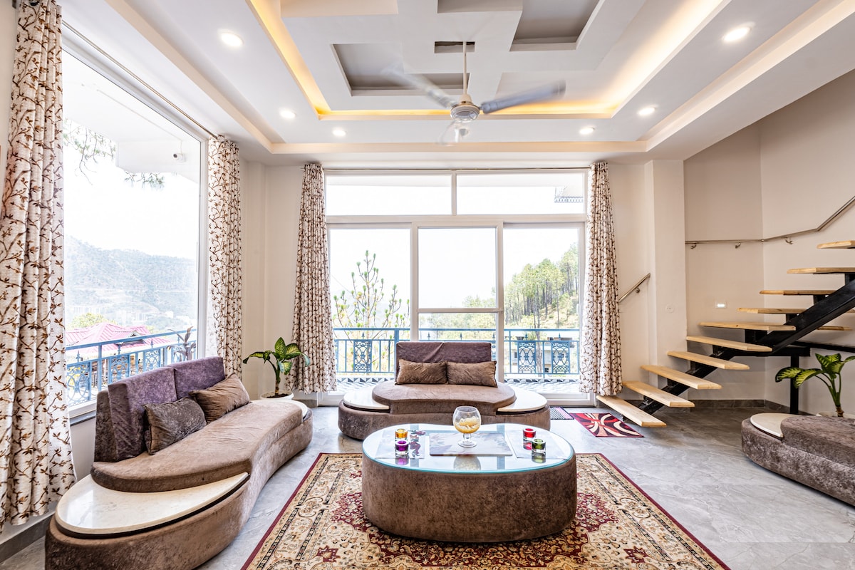 The spacious living room features large windows that offer a view of the surrounding hills. Comfortable seating is arranged around a central glass table, while a decorative rug adds warmth to the space. A stylish staircase leads to the upper level.