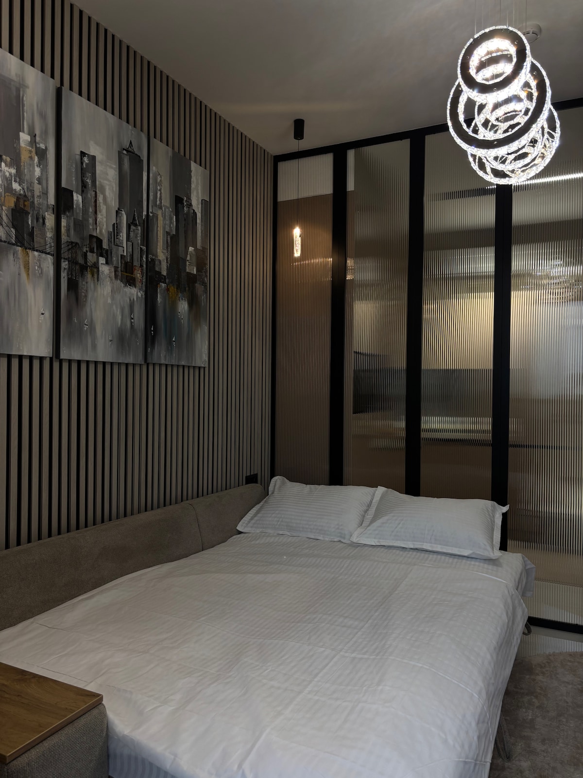 A modern sleeping area features a spacious bed with crisp white linens. A unique chandelier provides overhead lighting, while a contemporary wall art piece depicting a cityscape adds visual interest. Floor-to-ceiling glass panels separate the space, enhancing the open feel.