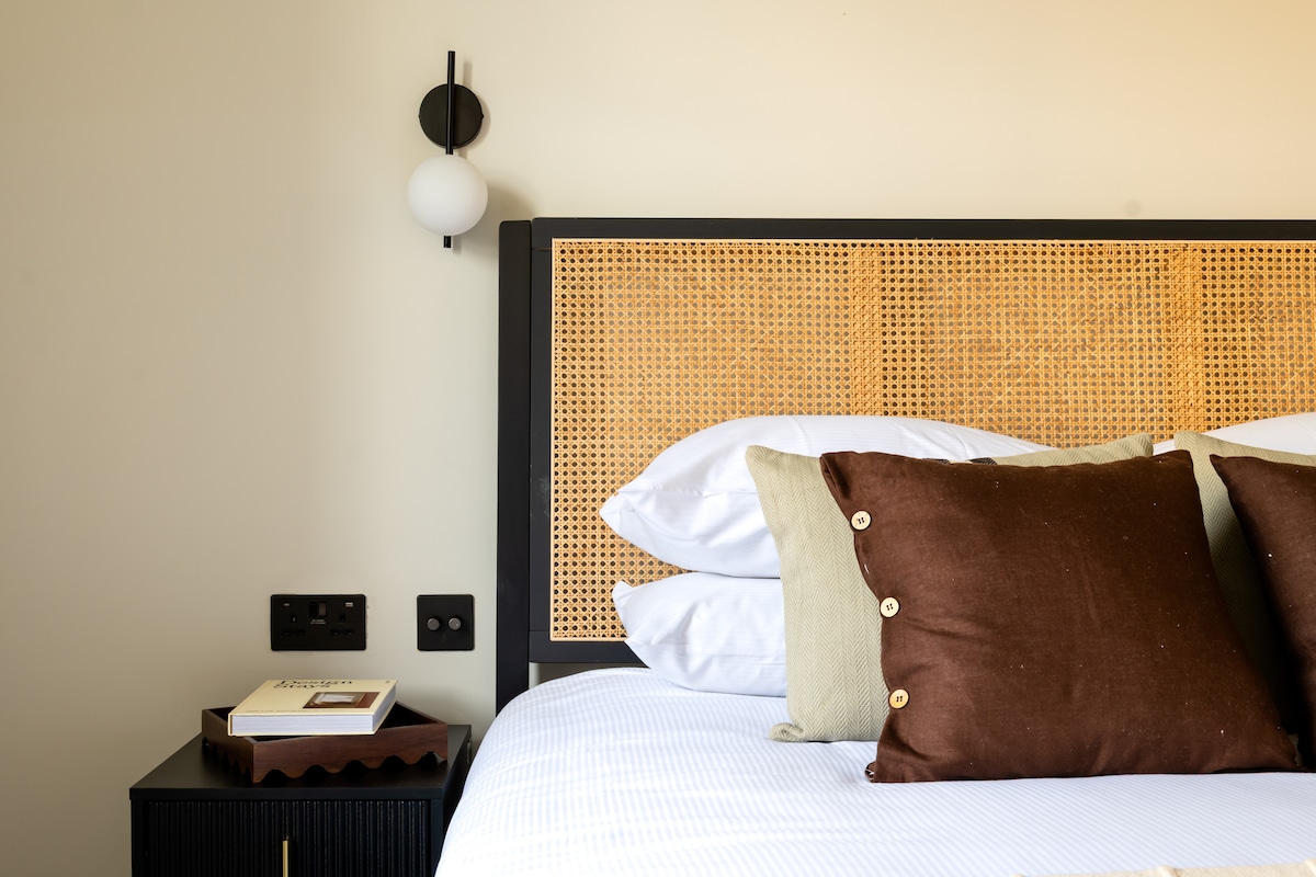 A stylish sleeping area features a woven headboard with soft, neutral-toned bedding. Cushions in various shades add texture. A bedside table displays a book, and a modern light fixture is mounted on the wall, contributing to the contemporary design.