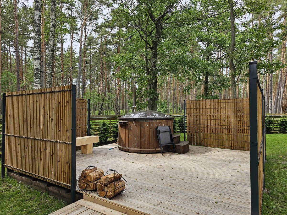 A spacious wooden deck features a round hot tub surrounded by natural scenery. Tall trees are visible in the background, providing a serene setting. Nearby, a wooden bench and a single chair are positioned for relaxation, while neatly stacked firewood adds a rustic touch.