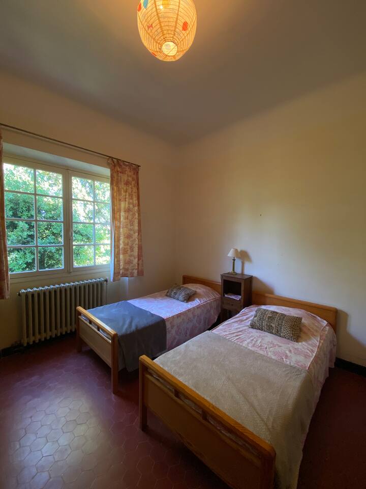 Une chambre épurée avec deux lits simples, parfaite pour accueillir petits et grands. La fenêtre offre une belle vue sur le jardin arboré et laisse entrer la lumière du matin.