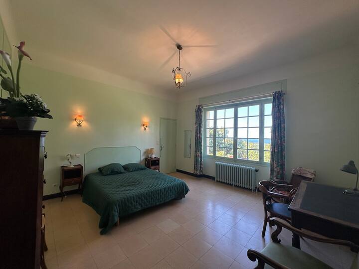 Une grande chambre avec lit double, parquet clair et fenêtres ouvertes sur la verdure. Atmosphère paisible, parfaite pour des nuits reposantes dans un cadre naturel.