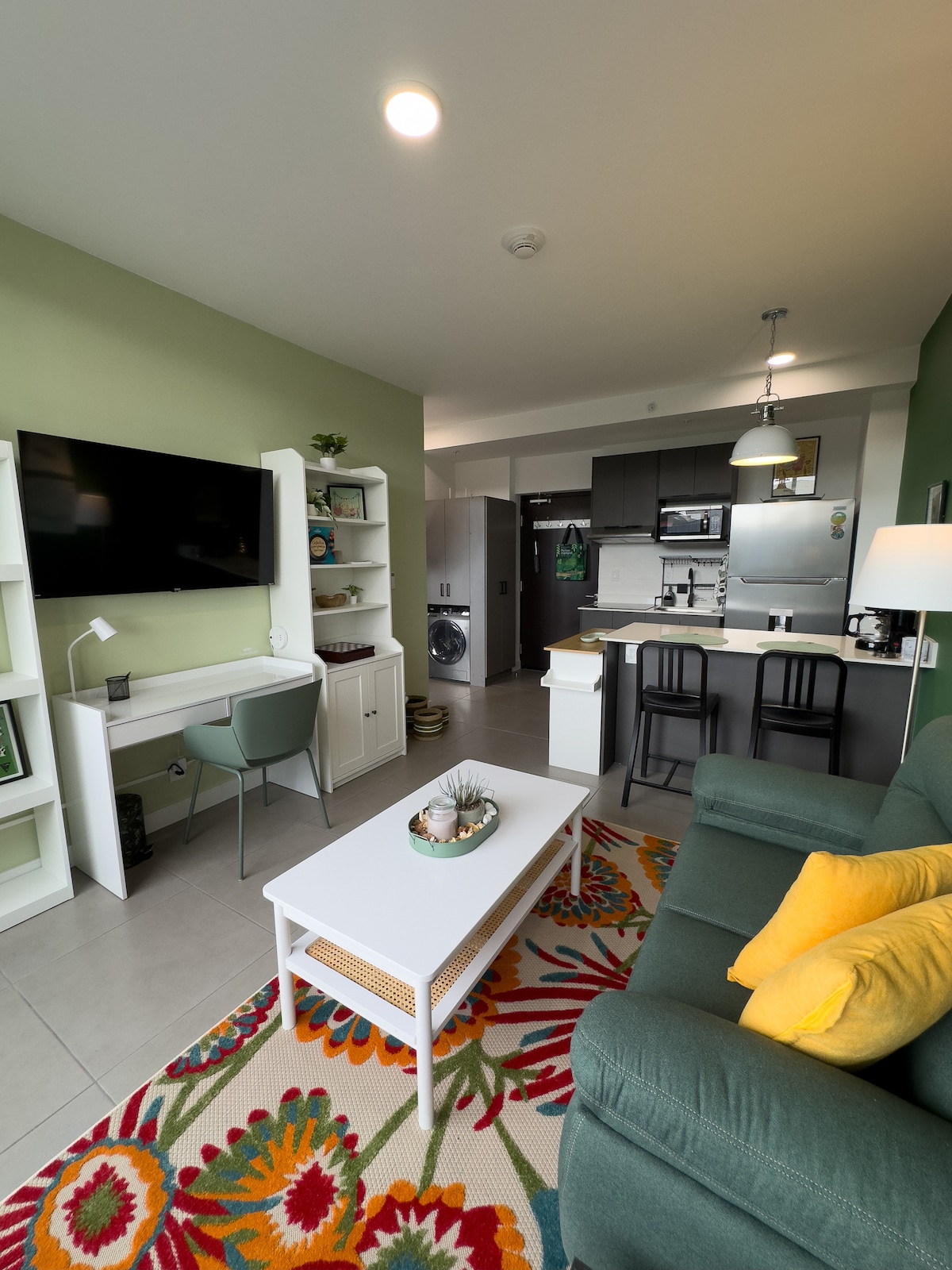 A modern living area is showcased, featuring a comfortable green sofa adorned with yellow cushions. A coffee table sits centrally, accompanied by a vibrant rug with floral patterns. A wall-mounted TV is visible, alongside a small dining space and a kitchen equipped with stainless-steel appliances.