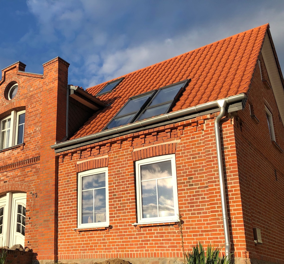 A traditional brick building is shown featuring a bright orange tile roof. Two large windows reflect the sky, and solar panels are installed on the sloped roof. Greenery is visible at the base of the structure, emphasizing its classic architectural style.