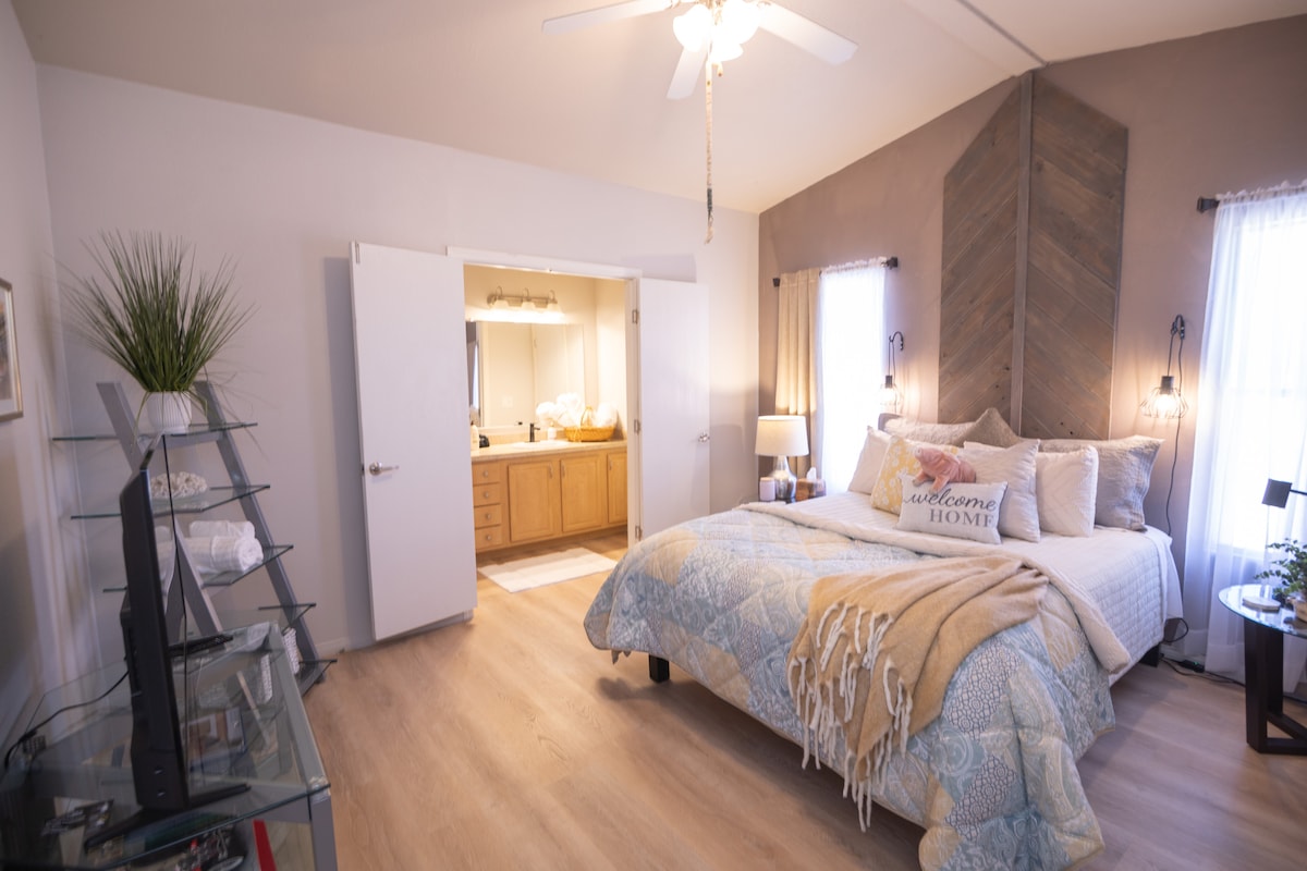 A bright bedroom features a spacious bed adorned with decorative pillows and a soft blanket, complemented by stylish nightstands. The room includes a ceiling fan and a large window with flowing curtains. An ensuite bathroom is accessible through double doors.