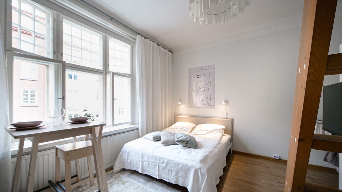 A bright sleeping area features a spacious bed dressed in crisp white bedding, complemented by soft green pillows. Natural light floods through large windows adorned with white curtains. A small dining table with two place settings is positioned nearby, enhancing the open feel of the room.