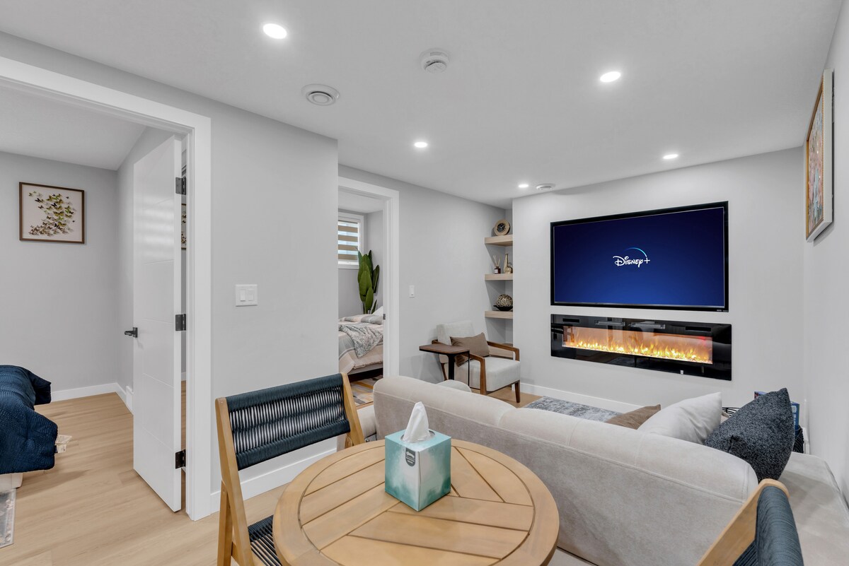 A modern living space is highlighted by a flat-screen TV mounted above a stylish electric fireplace. Soft seating is arranged around a round wooden table, with a cozy blanket draped over a nearby bed in the background. Bright lighting enhances the welcoming ambiance.
