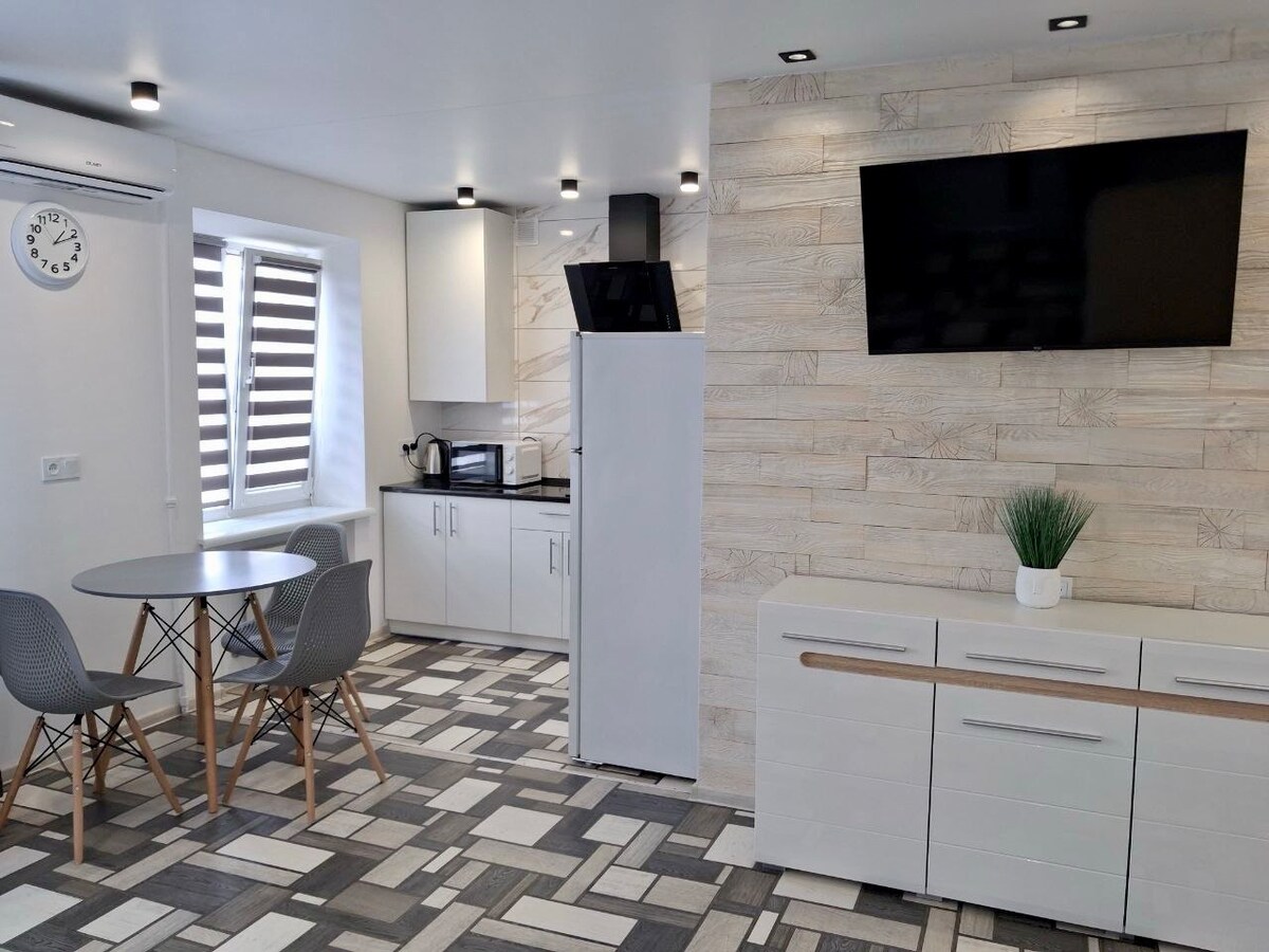 A modern kitchen area features a round dining table with gray chairs, adjacent to sleek white cabinetry. A large fridge and a microwave are visible, alongside a flat-screen TV mounted on a light wood-paneled wall. Soft lighting highlights the contemporary tile floor design.