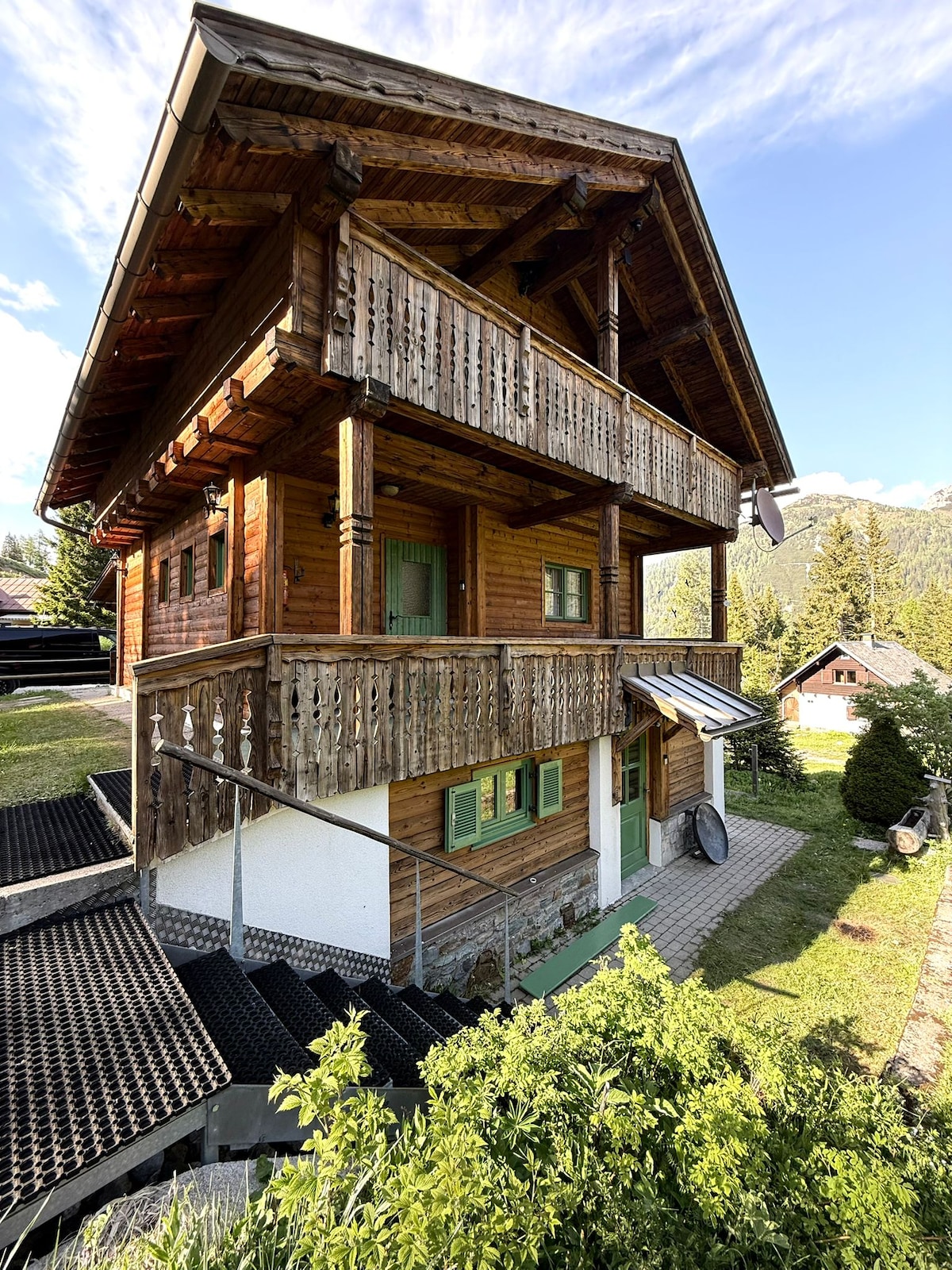 A rustic wooden chalet stands against a backdrop of blue skies and mountains, featuring a three-story structure with balconies and green shutters. A set of steps leads to the entrance, while landscaped greenery surrounds the base, enhancing the natural setting.