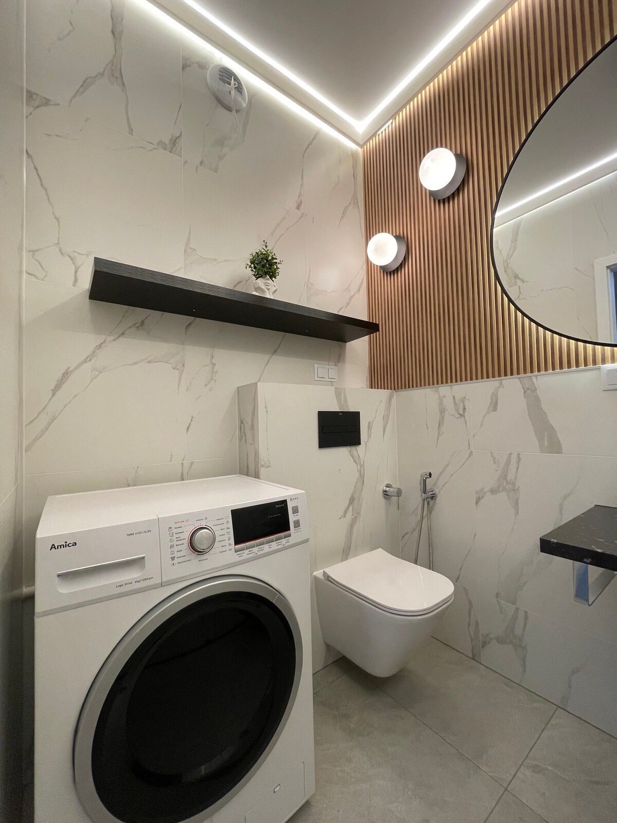 A modern bathroom features a washing machine positioned beside a wall-mounted toilet. Natural light is provided by a round mirror above a sleek sink, complemented by minimalist wooden accents. Soft LED lighting highlights the elegant marble walls and the clean, spacious design.