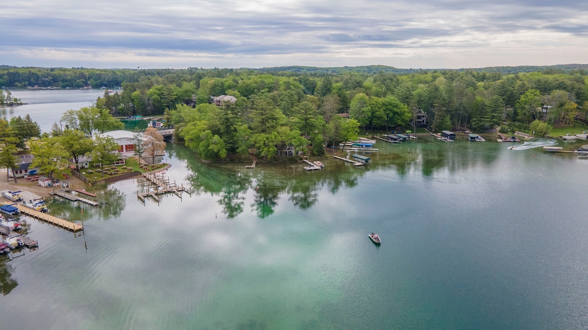An aerial view captures the serene Waupaca Chain of Lakes, showcasing lush greenery lining the shores. Small boats are visible on the gentle water, reflecting the surrounding landscape. Various docks and properties can be seen, highlighting the tranquil charm of the area.