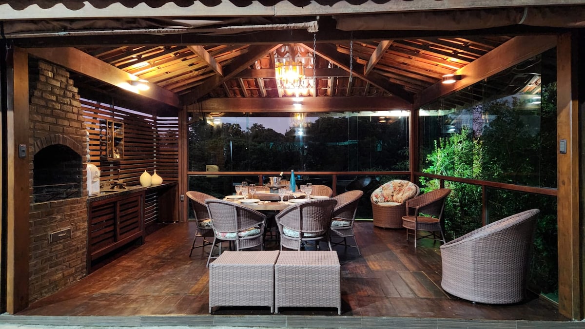 A spacious covered terrace features a wooden ceiling with decorative lighting. Comfortable seating is provided by woven chairs, and a dining table is set for a meal. A brick barbecue grill is positioned nearby, with greenery visible through large glass panels.