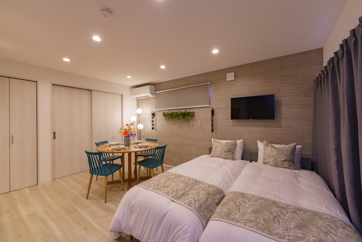 A bright, modern room features two spacious beds with soft linens and decorative pillows. A small dining table with teal chairs sits nearby, complemented by fresh flowers. Ambient lighting highlights the natural tones of the wood accents and the elegant wall-mounted TV.