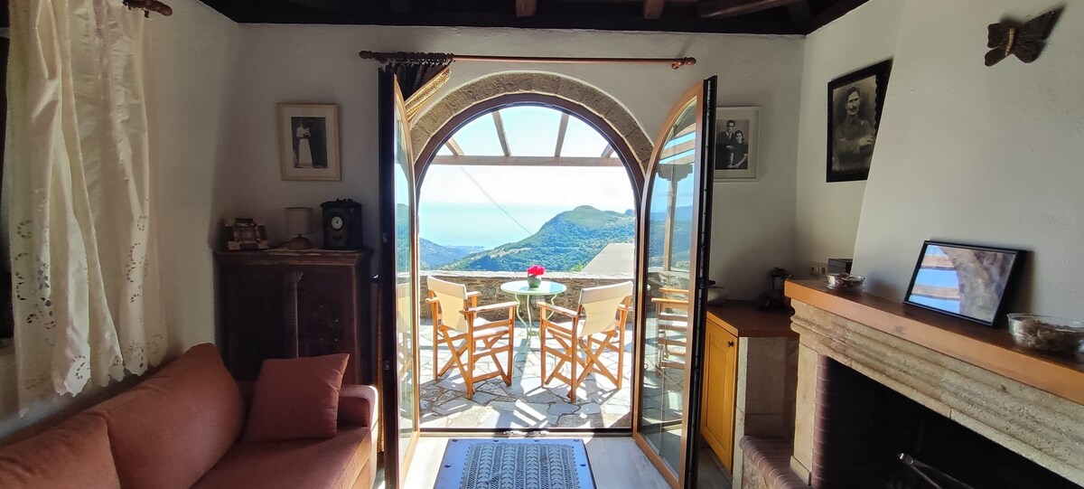 A cozy living area features an open doorway leading to a balcony with a view of the sea. Two wooden chairs and a small table are positioned outside, inviting relaxation. Natural light filters in through the doorway, illuminating the room's simple decor and soft furnishings.
