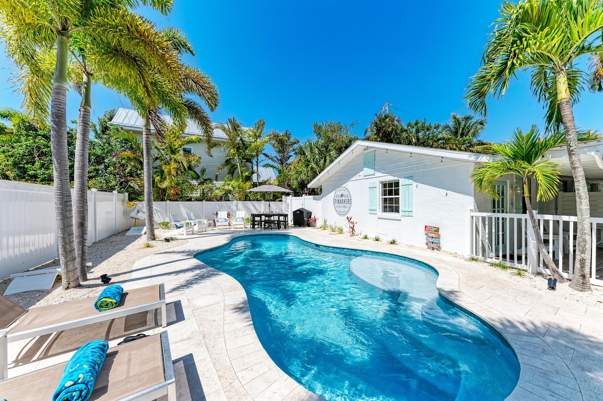 A tranquil outdoor area features a swimming pool surrounded by lush palm trees and tropical plants. Two loungers invite relaxation beside the water, and a dining table is set for outdoor meals. Sunshine brightens the scene, enhancing the inviting ambiance.