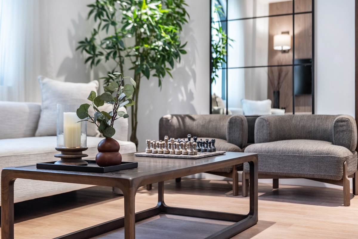 A modern living area features a light-colored sofa and a compact armchair. A wooden coffee table is adorned with a decorative vase and a chess set. Reflections can be seen in a large wall mirror, enhancing the space's openness.