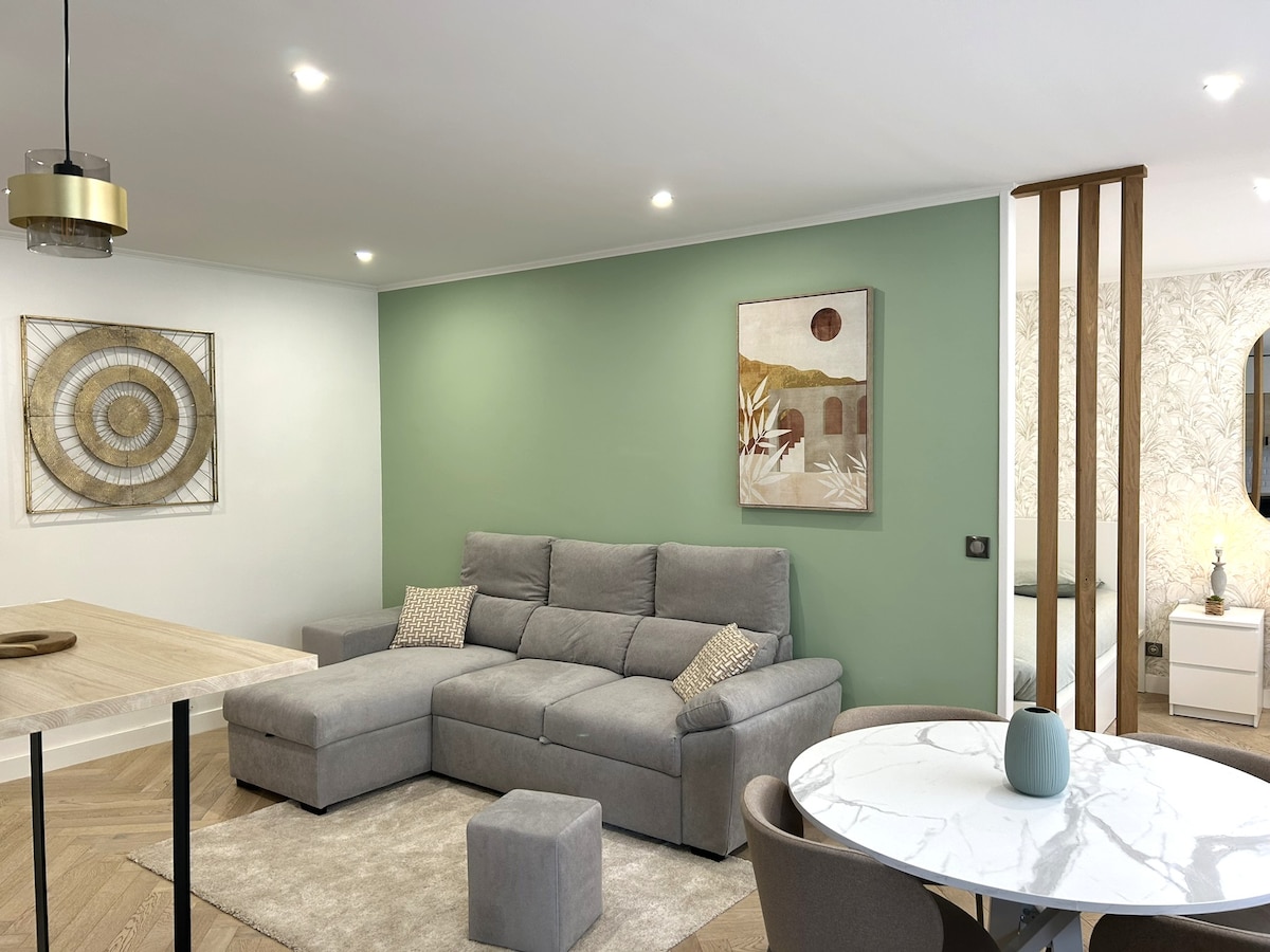 A modern living area features a grey sectional sofa with decorative pillows against a soft green accent wall. A marble dining table is set with round chairs, while a piece of artwork complements the decor. Soft lighting from recessed fixtures illuminates the space.
