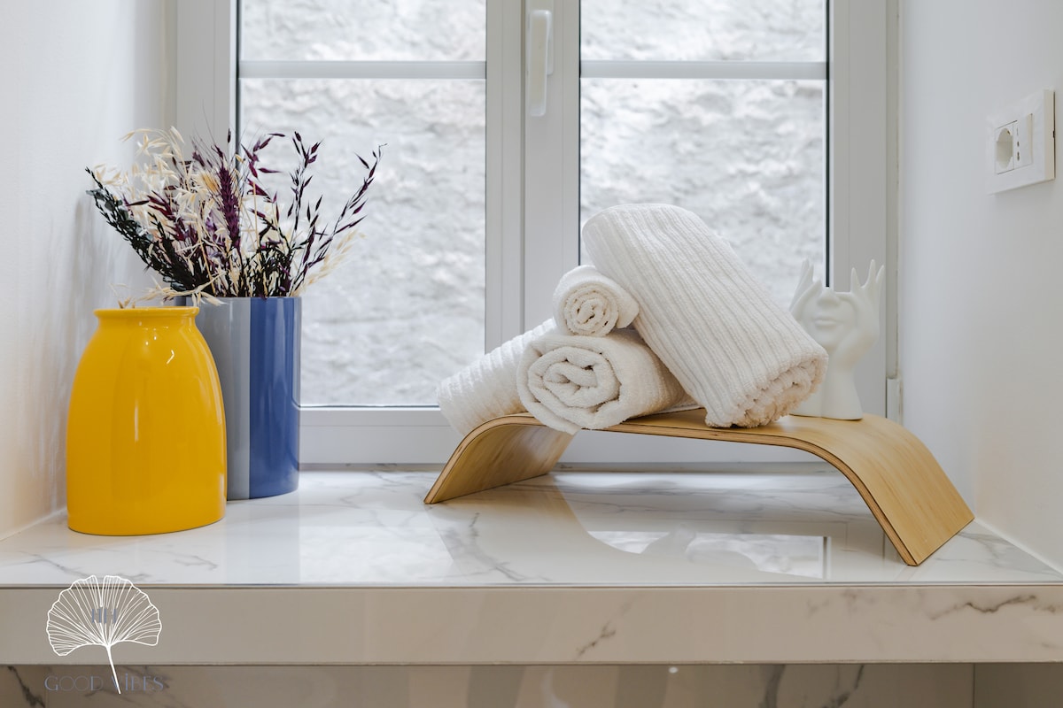 A bright countertop displays a bamboo towel holder, neatly stacked with rolled towels. A yellow vase holds dried flowers, complemented by a blue vase. Natural light filters through a window, enhancing the clean and modern aesthetic of the space.
