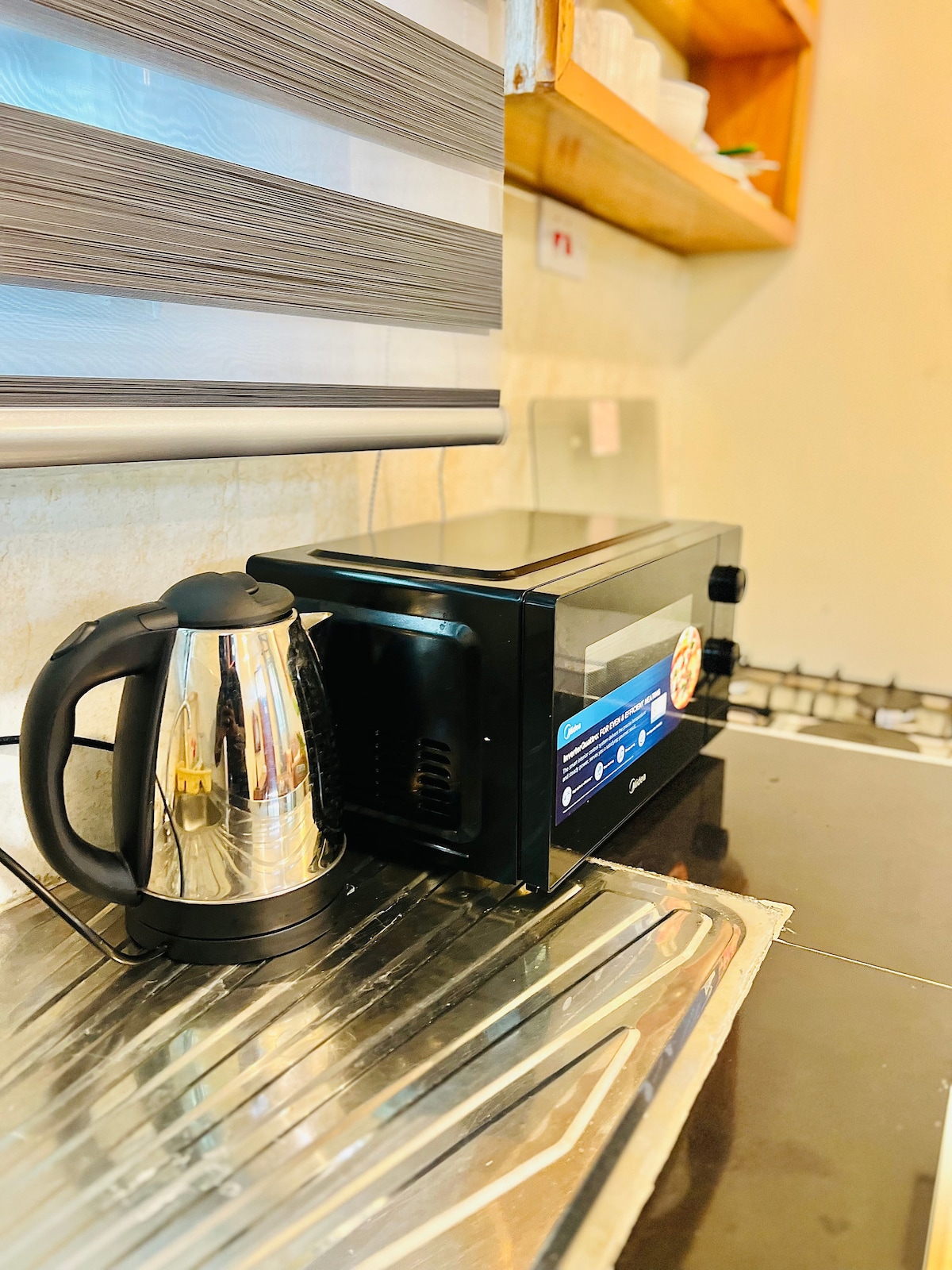 A modern kitchen countertop is presented, showcasing a sleek microwave and a stainless steel kettle. The countertop features a reflective surface, with additional kitchenware visible on the shelves above. Natural light filters through a nearby window, enhancing the clean and functional layout.