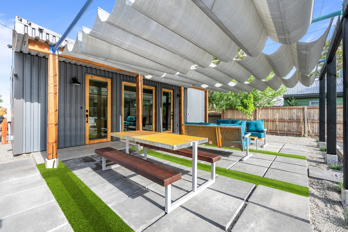 A spacious patio is showcased, featuring a large wooden dining table and benches under a stylized canopy. Colorful lounge furniture is positioned nearby, with green grass accents lining the concrete surface. The open area is surrounded by a fenced yard, creating an inviting outdoor setting.