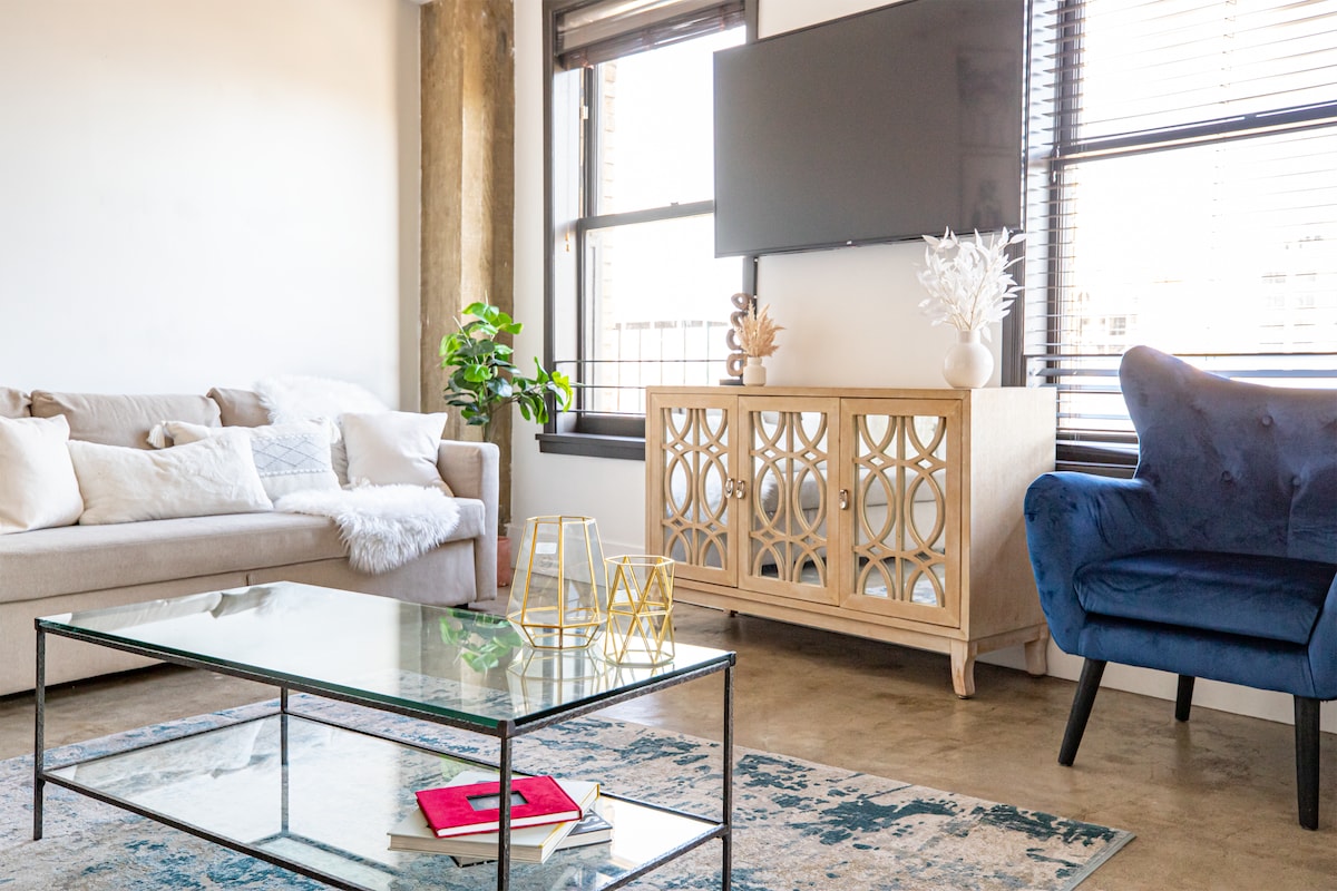 A modern living area is highlighted by a plush, light-colored sofa and a deep blue accent chair. A glass coffee table rests on a decorative area rug, with a media console featuring intricate designs. Natural light streams through large windows, creating a fresh and open ambiance.