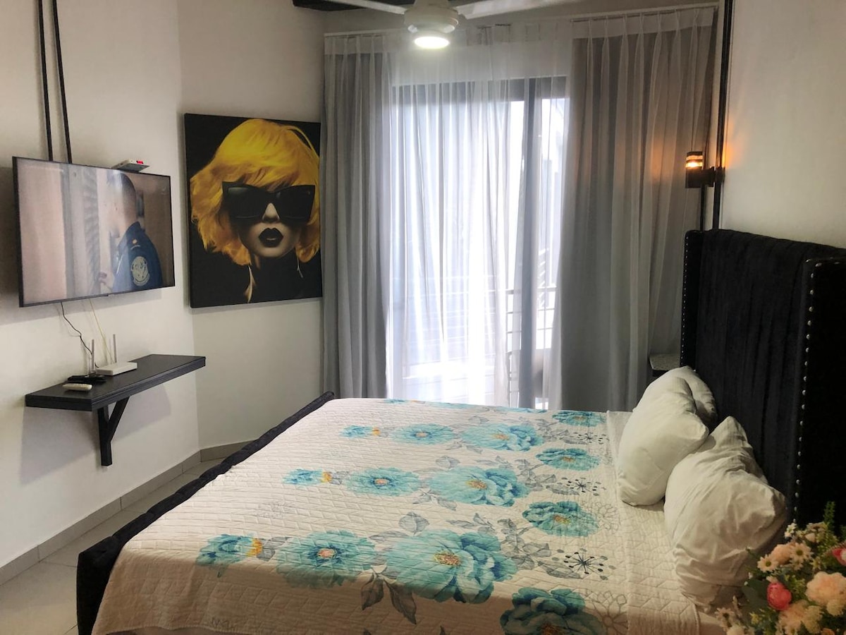 A comfortable sleeping area features a queen-size bed adorned with a floral patterned quilt and multiple pillows. A wall-mounted television is present, and large glass doors allow natural light to illuminate the space while providing access to a private balcony.