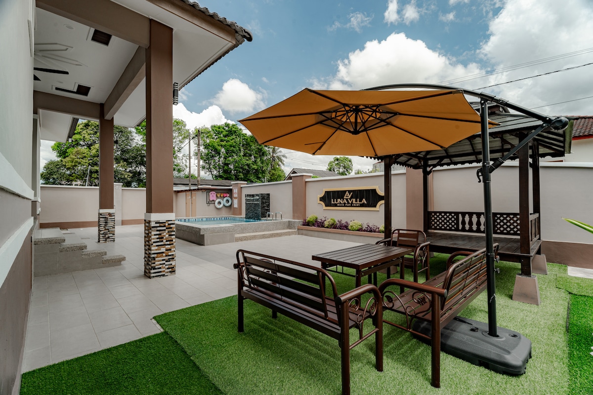 An outdoor seating area features a large shaded umbrella over a wooden table nestled among green grass. Nearby, a sturdy gazebo complements the space, while a private pool is visible in the background. The surroundings exhibit a well-maintained and relaxing environment.