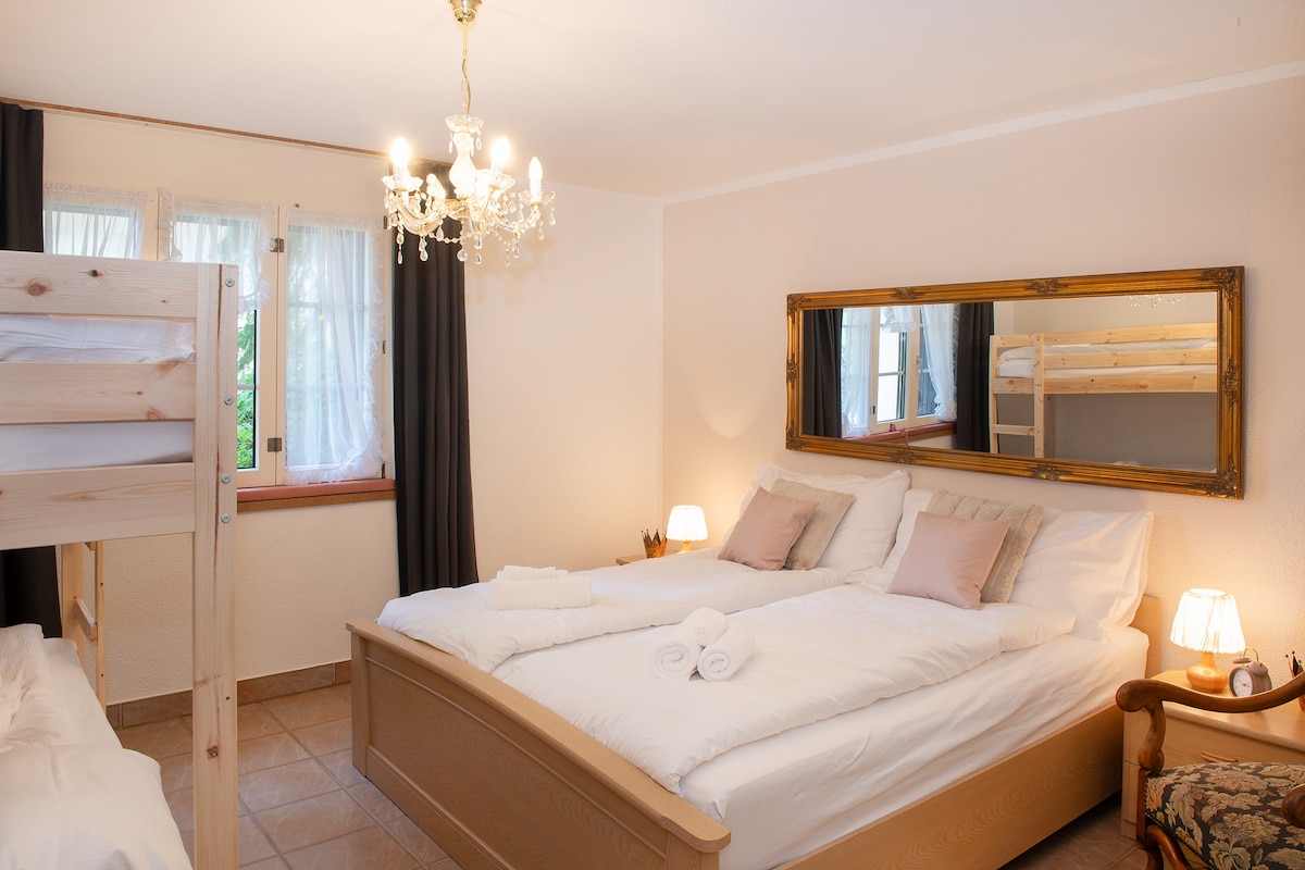 A bright bedroom features a spacious double bed and a bunk bed against one wall. Soft linens and pillows are neatly arranged. A large mirror reflects the room's warm tones, while bedside lamps and a vintage armchair create a cozy ambiance. Windows allow natural light to fill the space.