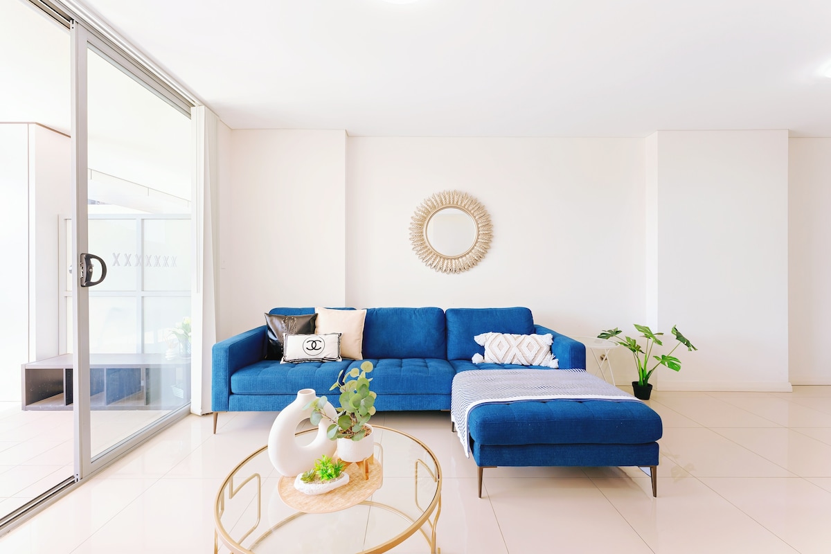 A modern living area is depicted, featuring a blue sectional sofa accompanied by a decorative throw blanket. A round coffee table with plants and a decorative vase is positioned in front. Natural light enters through large glass doors, highlighting a relaxed environment.