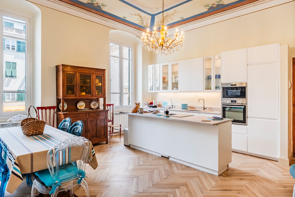 A spacious kitchen area features a central island with modern appliances and an elegant chandelier overhead. A large table is set for dining, complemented by vintage cabinetry. Decorative accents illuminate the high ceilings adorned with intricate frescoes.