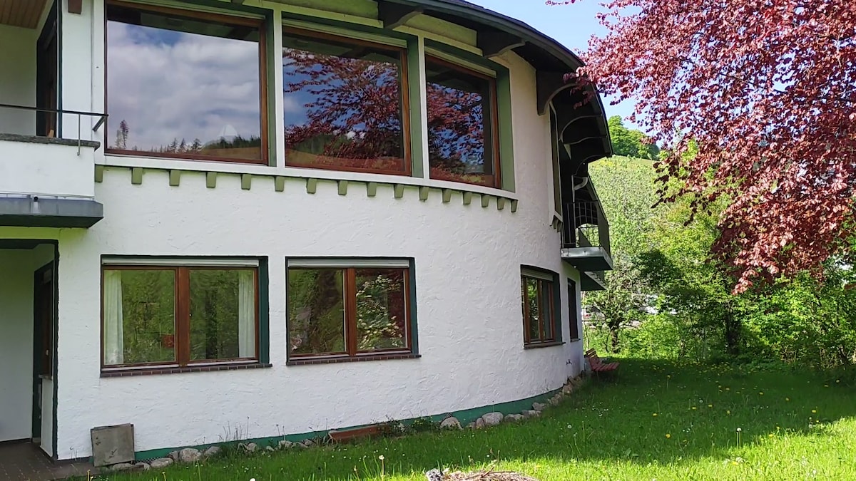 The exterior of the house showcases a softly rounded design and presents multiple large windows allowing natural light to enter. The vibrant green lawn surrounds the building, complemented by a flowering tree, providing a serene outdoor environment.